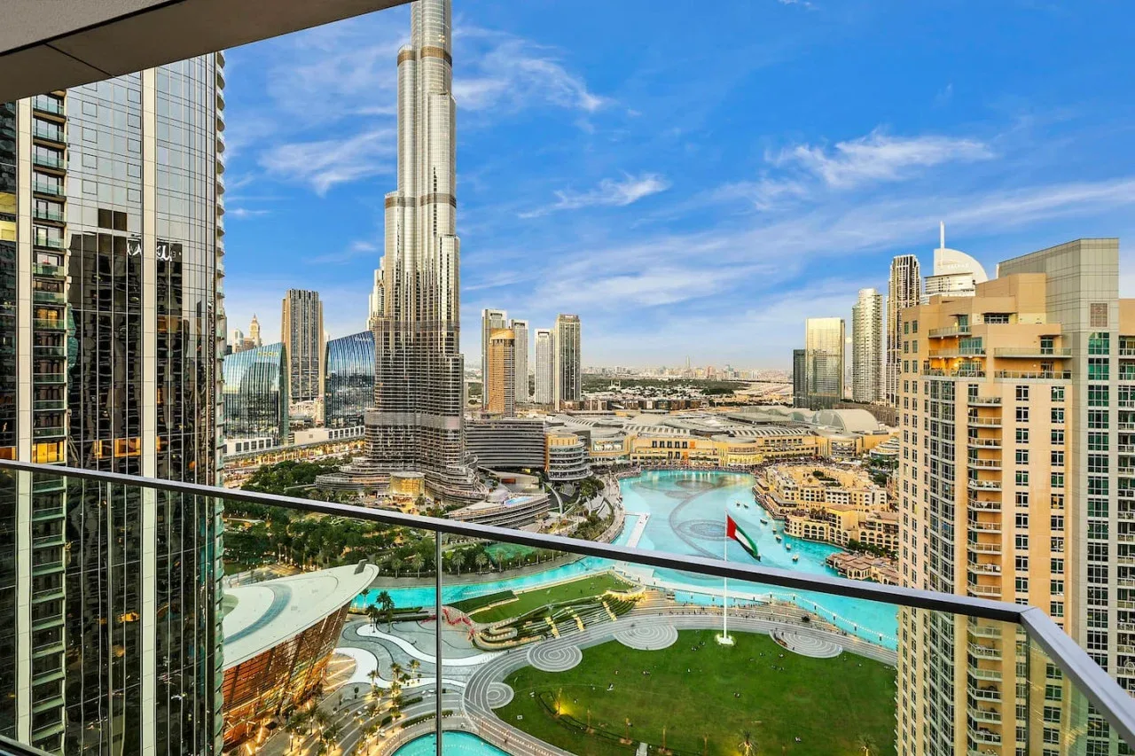A city skyline viewed from a balcony, with a tall central tower and landscaped grounds below.