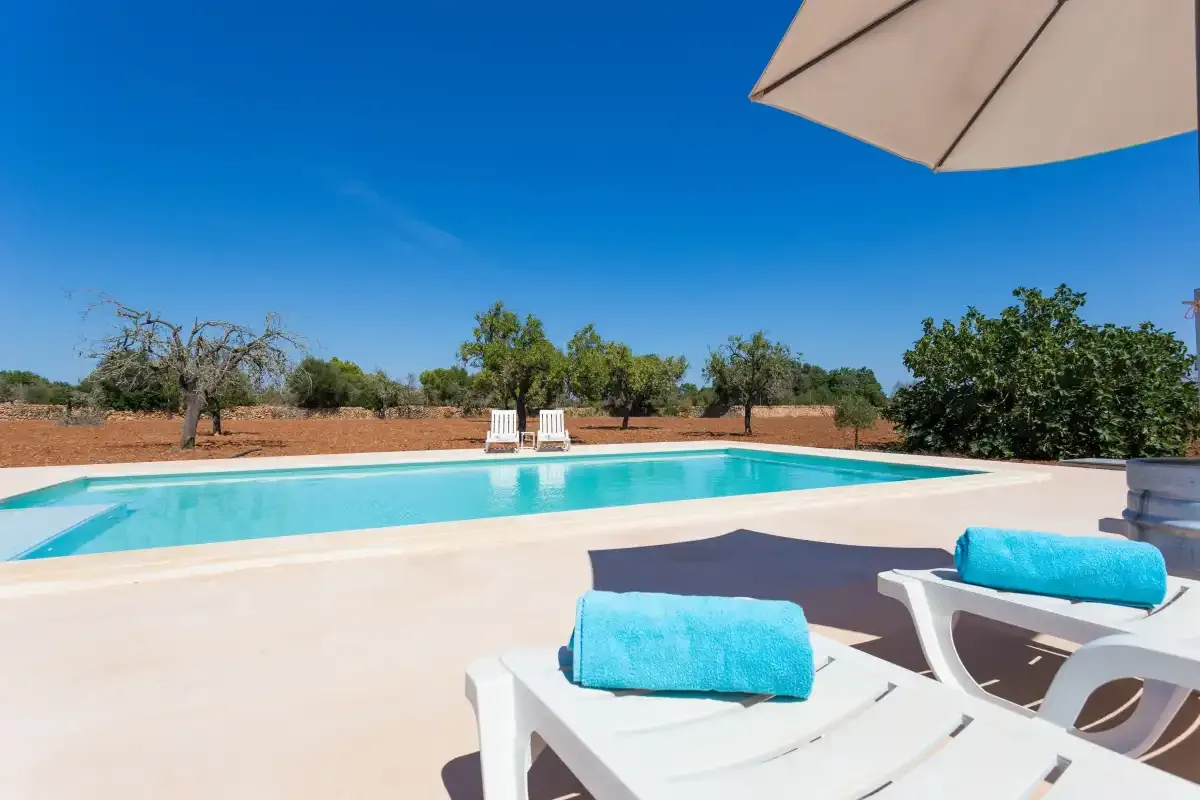 An outdoor swimming pool with loungers beside a dry, tree-lined landscape.