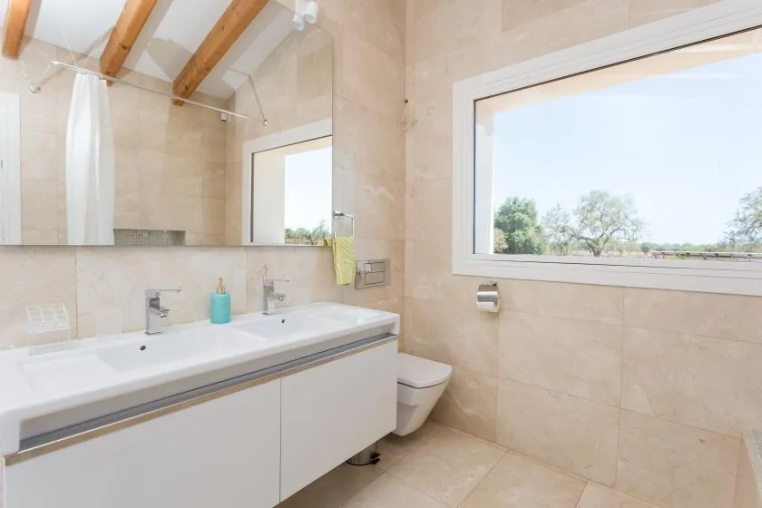 A bright tiled bathroom with a long sink counter and a large window.