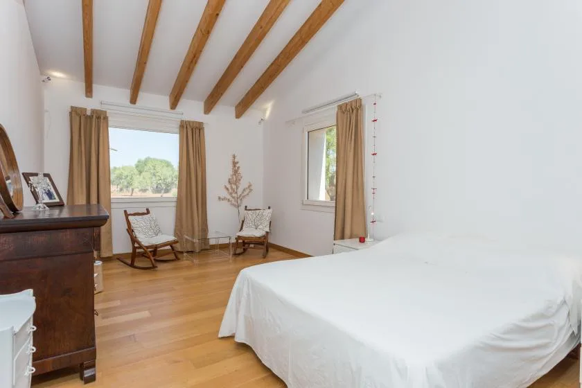 A bright bedroom with a white bed, wooden floor, and exposed ceiling beams.