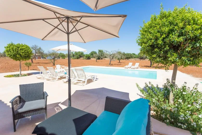 Un patio soleado junto a la piscina con asientos, sombrillas grandes y árboles frondosos.