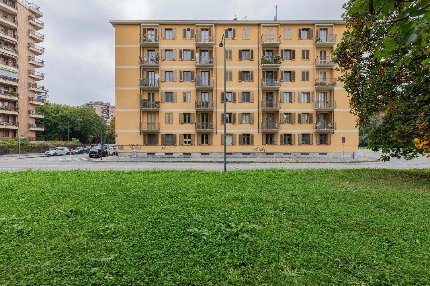 Un edificio de apartamentos se alza tras un césped, con árboles y edificios vecinos cerca.