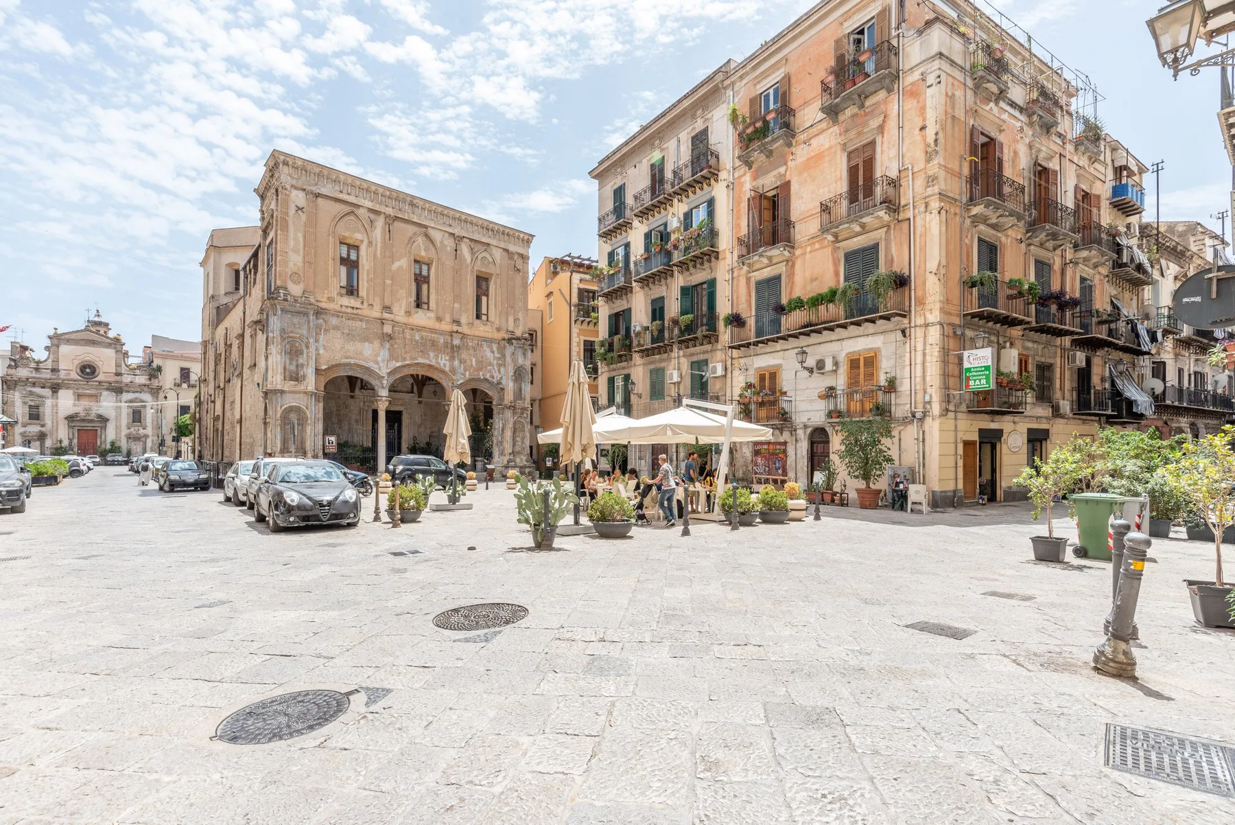 Una plaza pavimentada con edificios históricos, coches aparcados y mesas de café al aire libre.