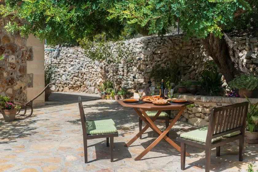 Una zona de comedor al aire libre con suelo de piedra, mesa y asientos bajo árboles frondosos.