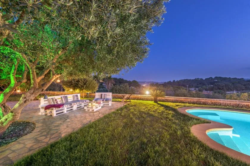 Un patio exterior con asientos da a una piscina y un paisaje arbolado al anochecer.