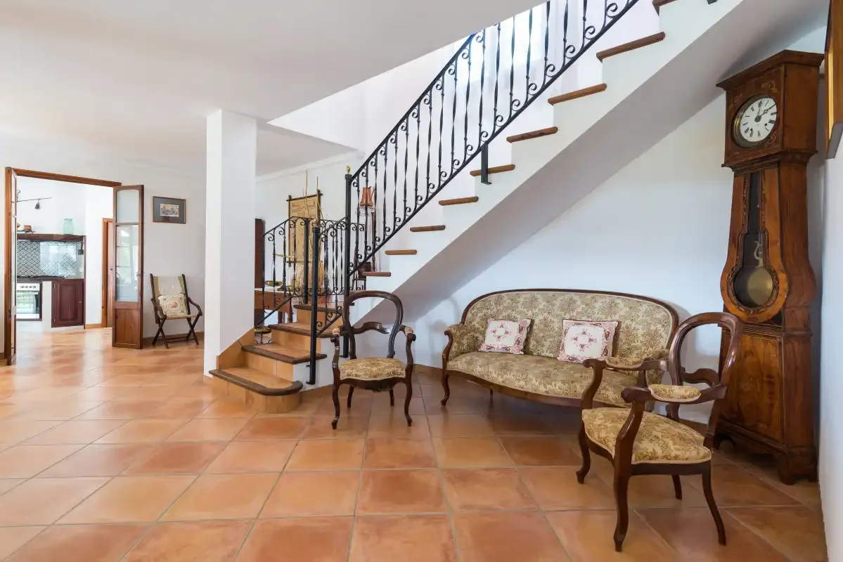 Una sala con suelo de baldosas, asientos bajo una escalera y un reloj alto de madera.