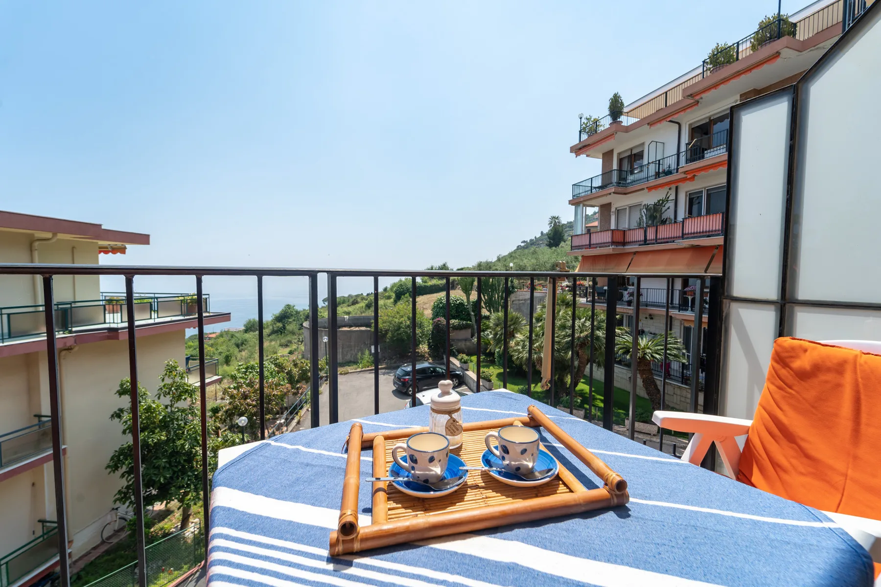 A balcony table holds a tray of cups beside orange seating, with buildings and greenery beyond.