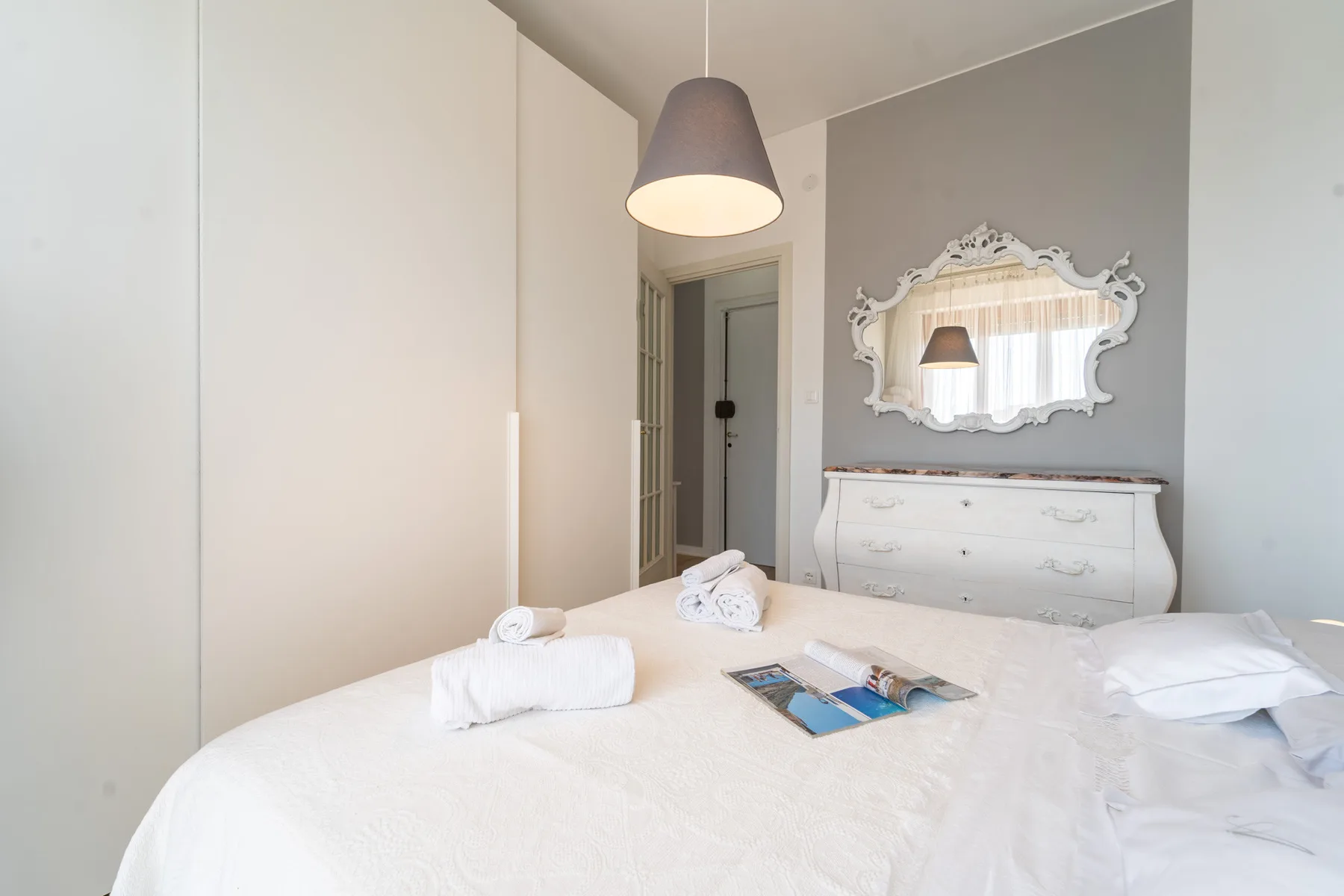 A bright bedroom with a white bed and a dresser beneath an ornate mirror.