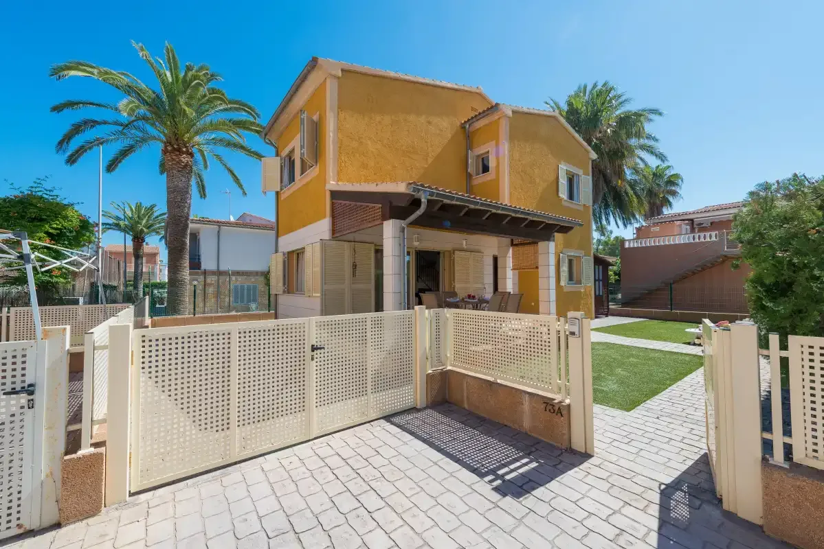 A yellow house stands behind a white fence beside a paved walkway.