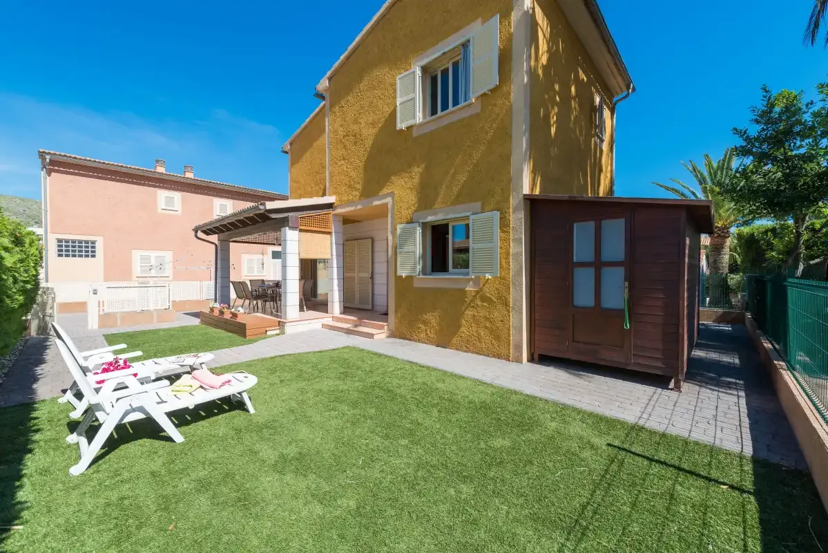 A house opens onto a small lawn with white lounge chairs beside it.