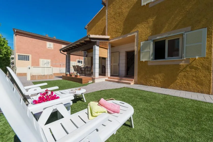 A sunny patio with white lounge seating beside a yellow house and green lawn.