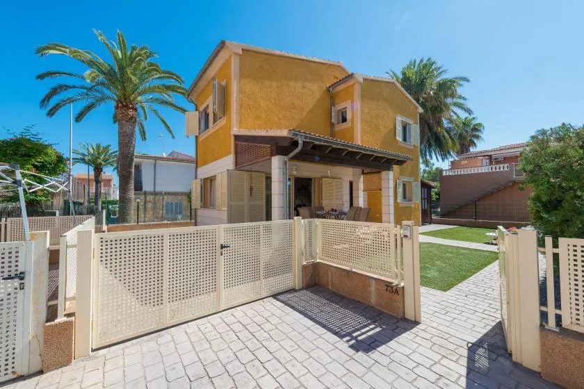 A yellow two-story house with a paved entrance, lawn, and palm trees.