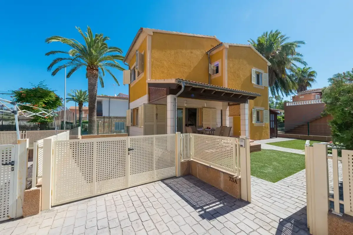 A yellow two-story house stands behind a paved entrance and white fencing.