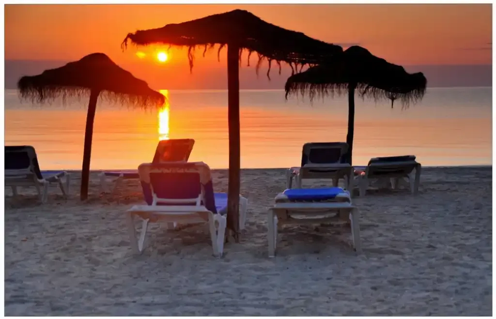 A beach at sunset with loungers beneath thatched parasols.