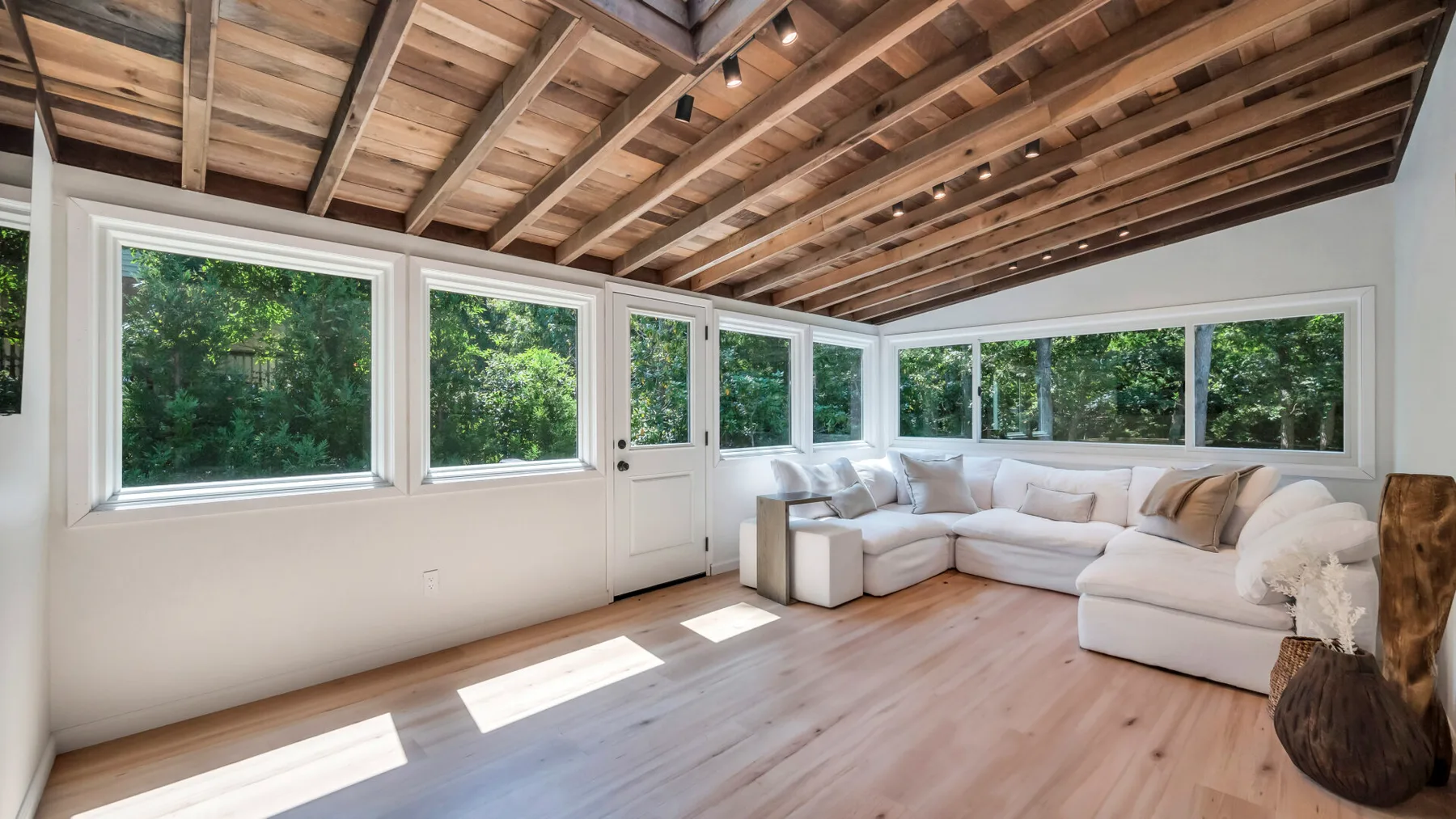 A bright enclosed sunroom with broad windows, leafy views, and a sectional seating area.