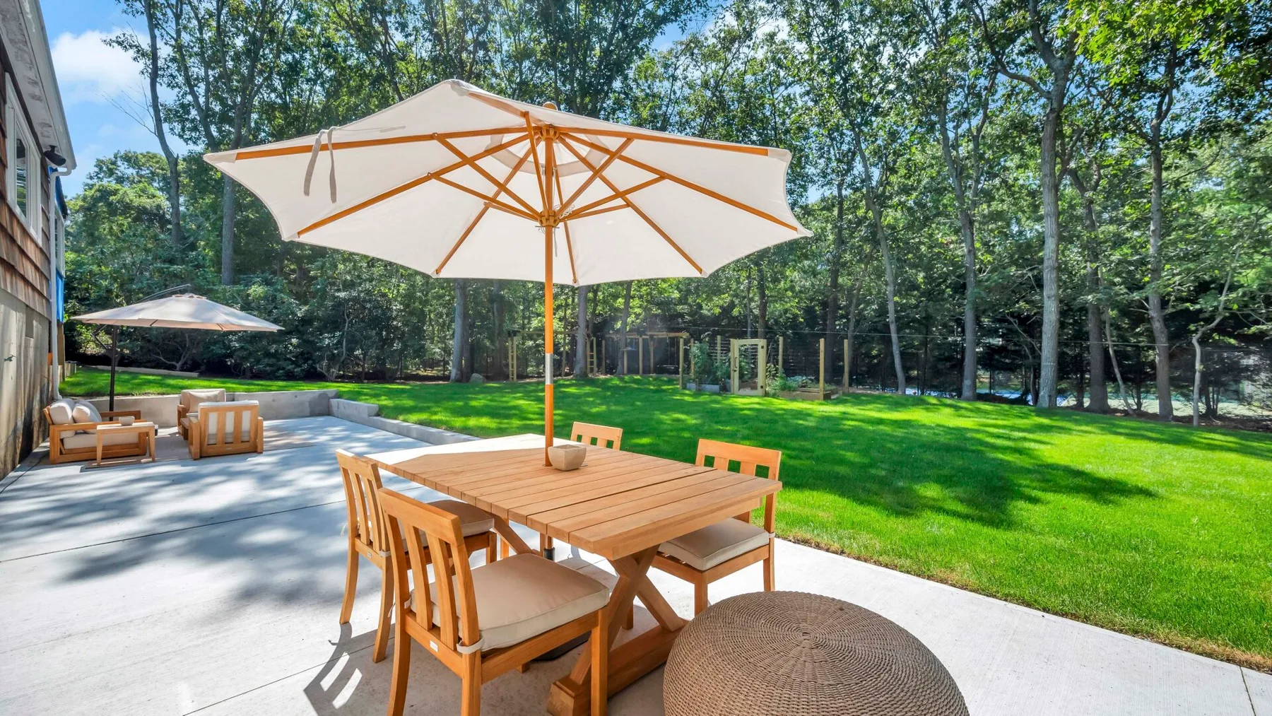 A wooden patio table and seating sit beneath an umbrella beside a green lawn.