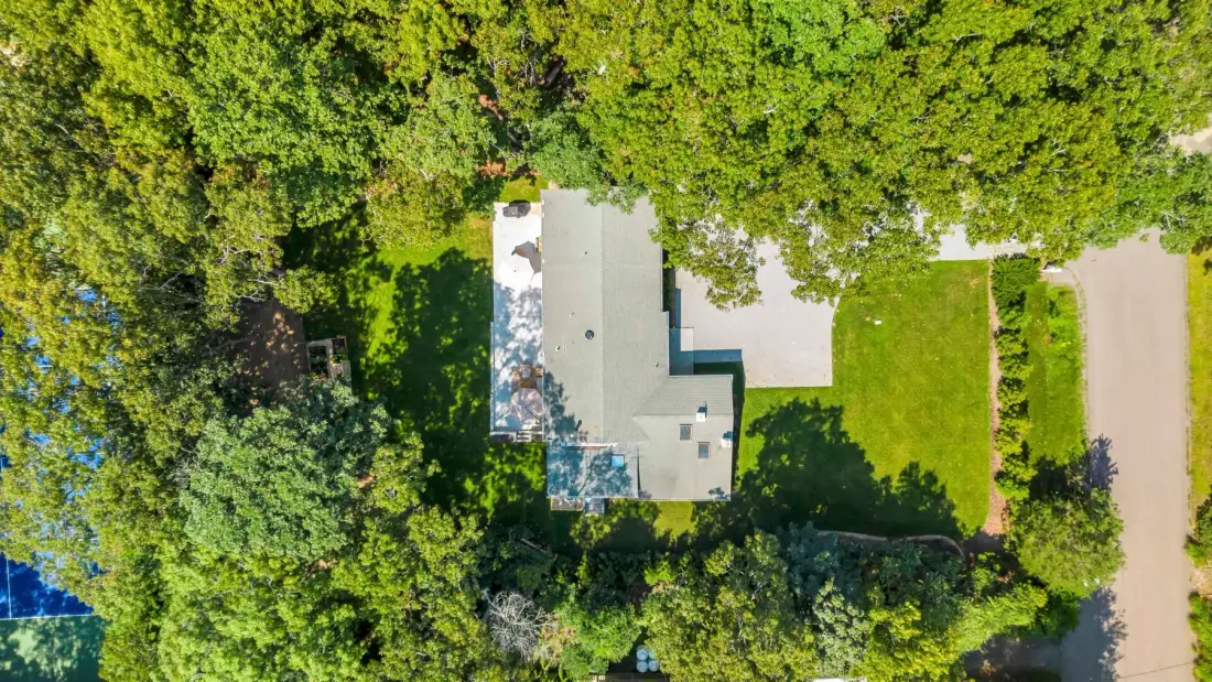 An aerial view shows a house surrounded by trees, with a lawn and driveway nearby.