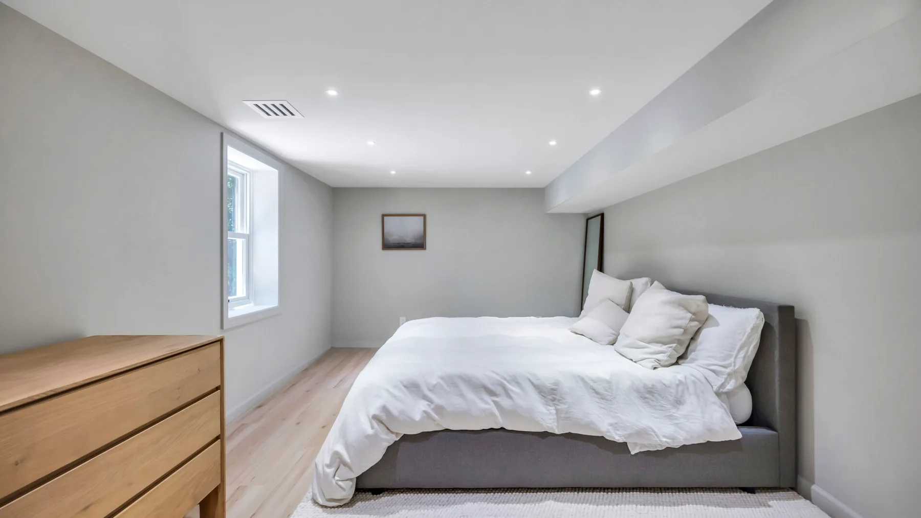 A bright bedroom with a bed dressed in white bedding and a wooden dresser.
