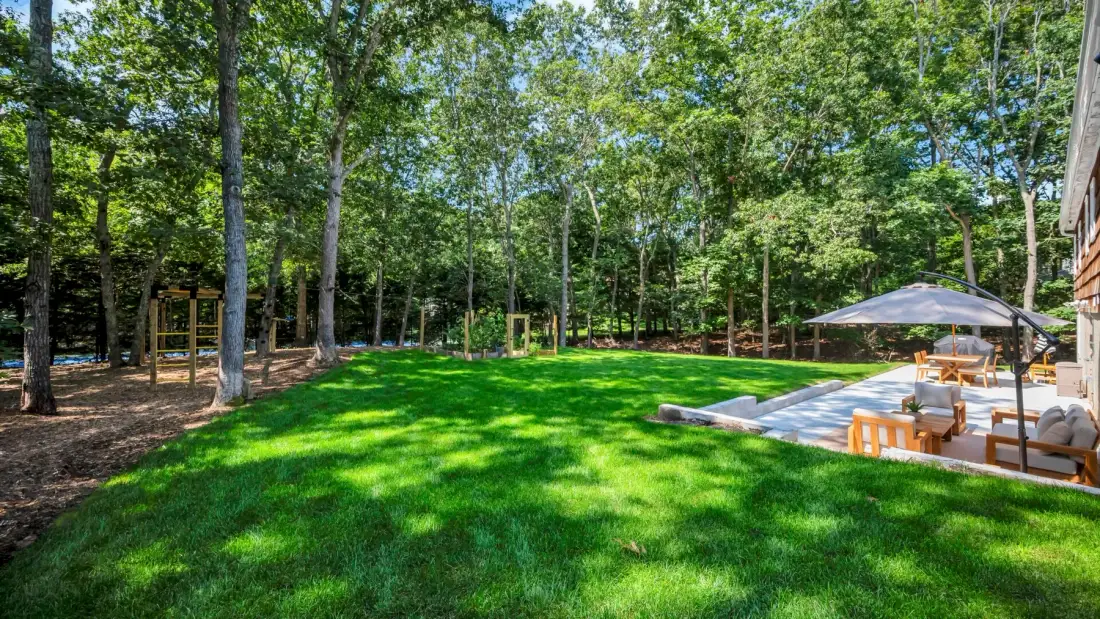 A grassy backyard bordered by trees, with outdoor seating beside a patio.