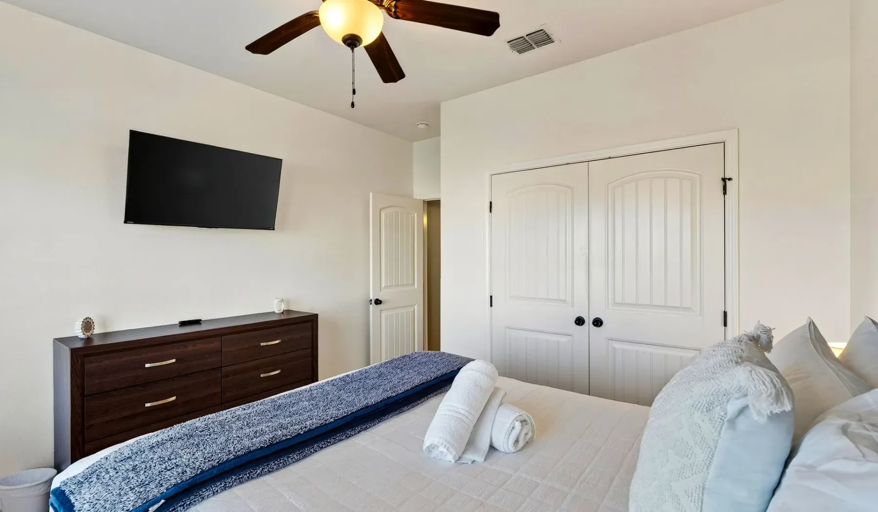 Un dormitorio con cama, cómoda oscura, televisor montado en la pared y ventilador de techo.