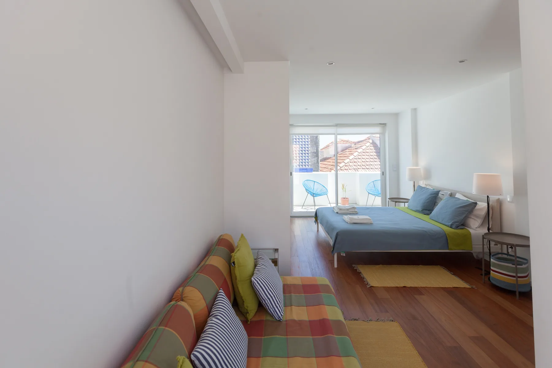 Un dormitorio luminoso con camas, asientos coloridos y una puerta de vidrio frente a tejados.