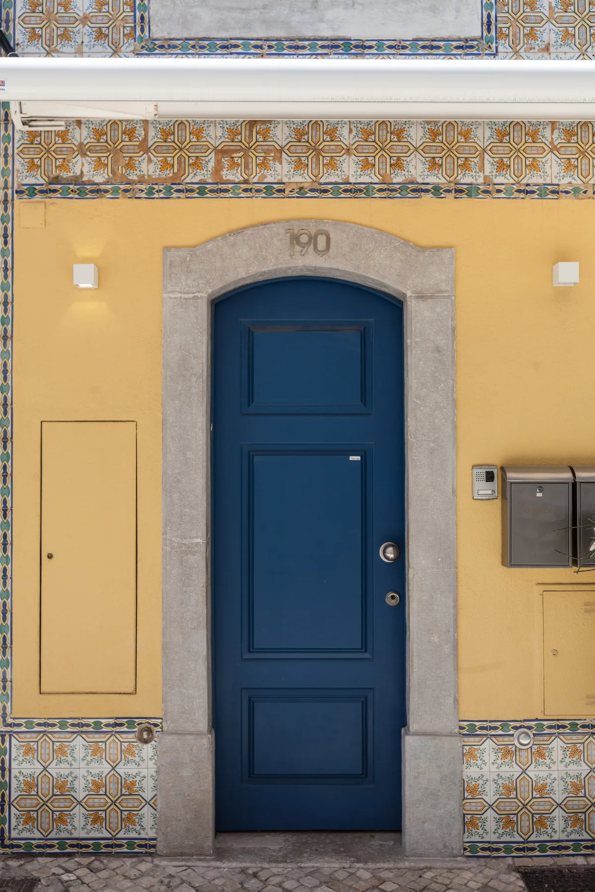 Una puerta azul de entrada se encuentra en una fachada amarilla con detalles de azulejos estampados.