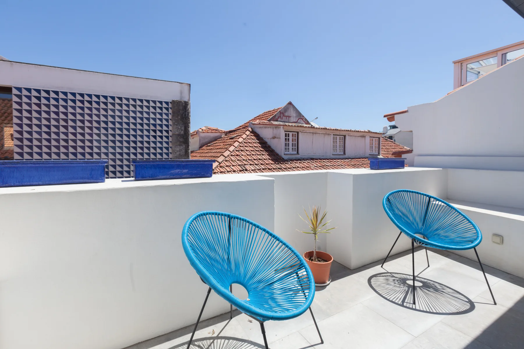 Una terraza en la azotea tiene asientos azules y una planta en maceta junto a un parapeto blanco.