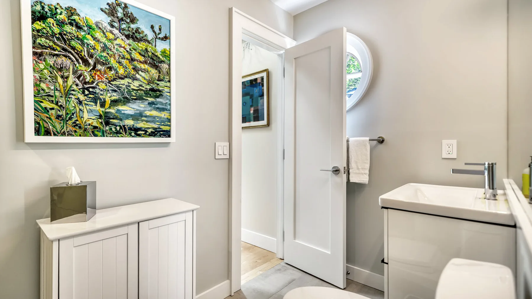 Un baño luminoso con lavabo, puerta abierta y una obra enmarcada.