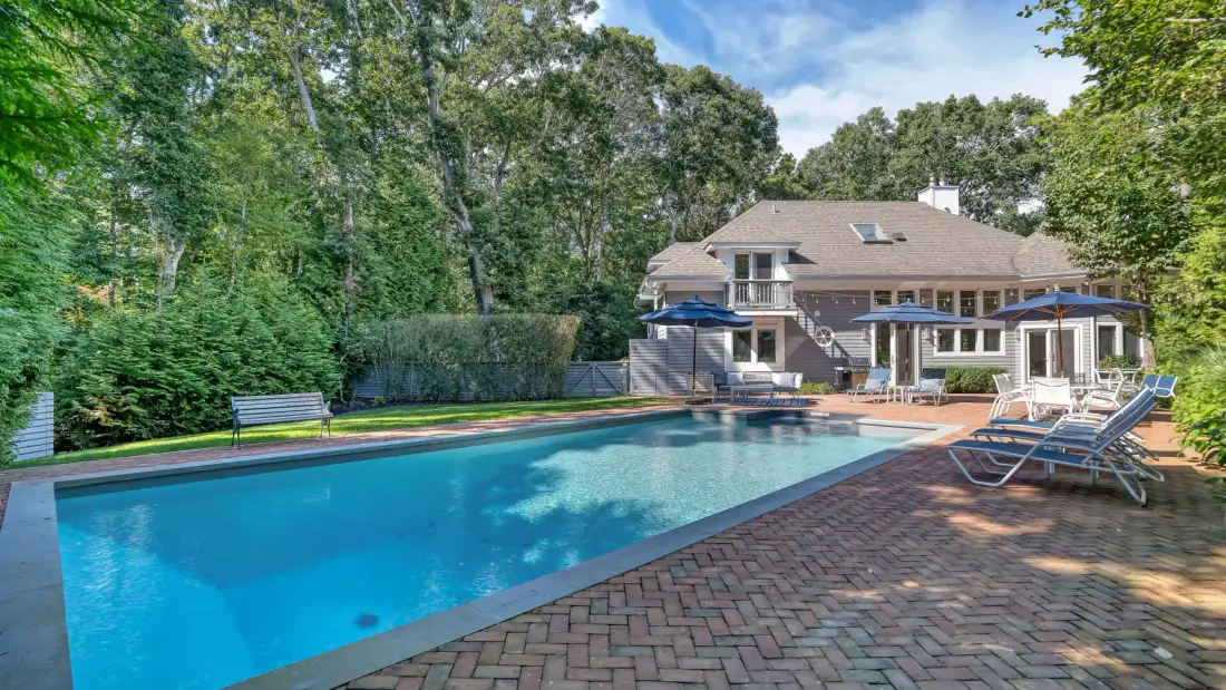 Una piscina en el patio trasero está junto a un patio pavimentado y una casa grande.
