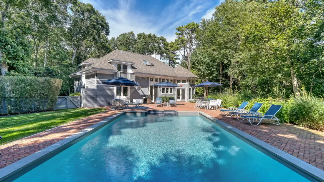 Una piscina y un patio están junto a una casa grande, rodeados de árboles frondosos.