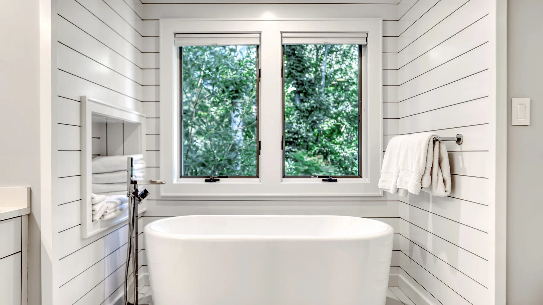 Un baño luminoso tiene una bañera exenta bajo dos ventanas con vista a árboles frondosos.