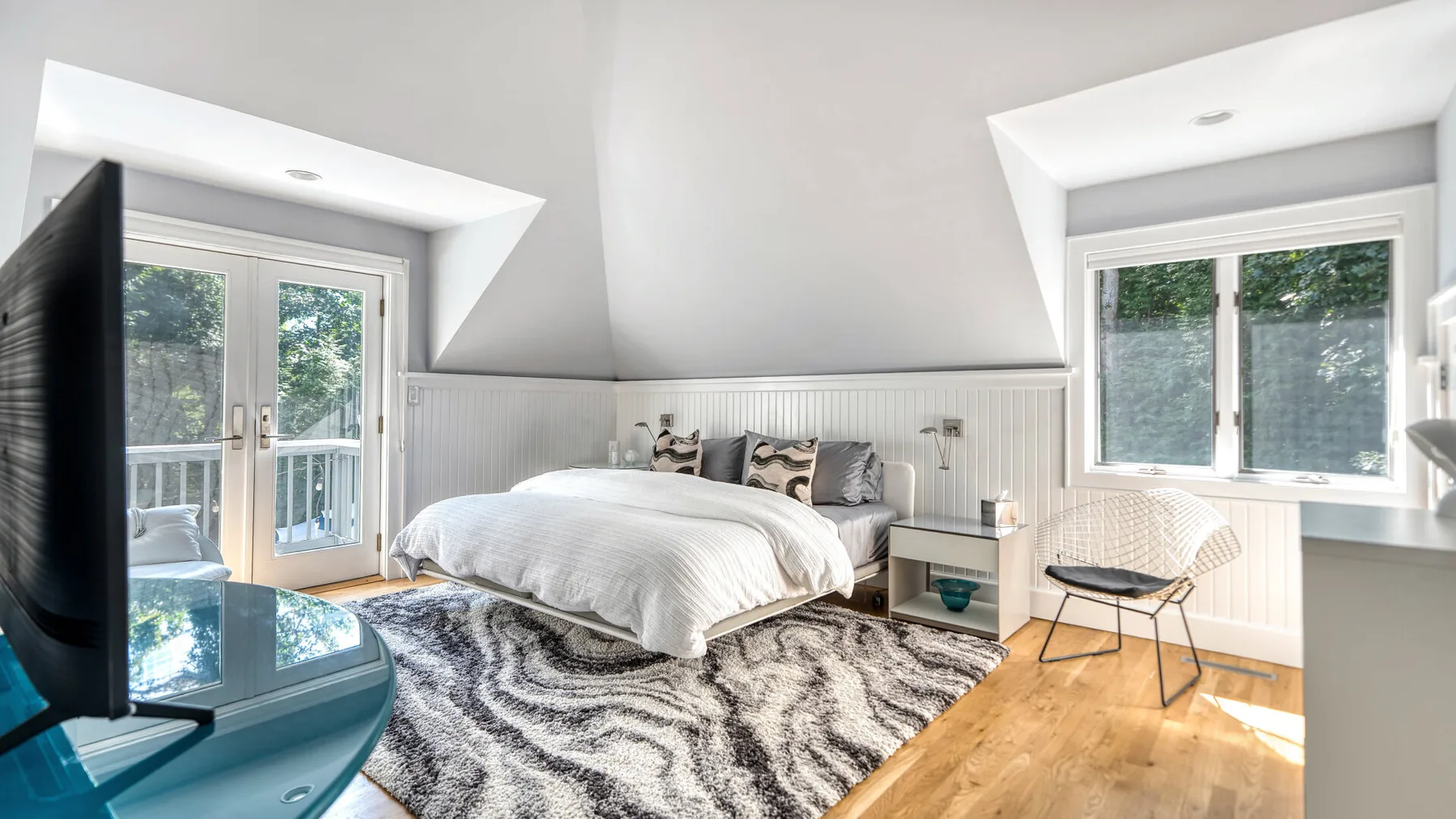 Un dormitorio luminoso con una cama, ventanas grandes y una alfombra estampada.