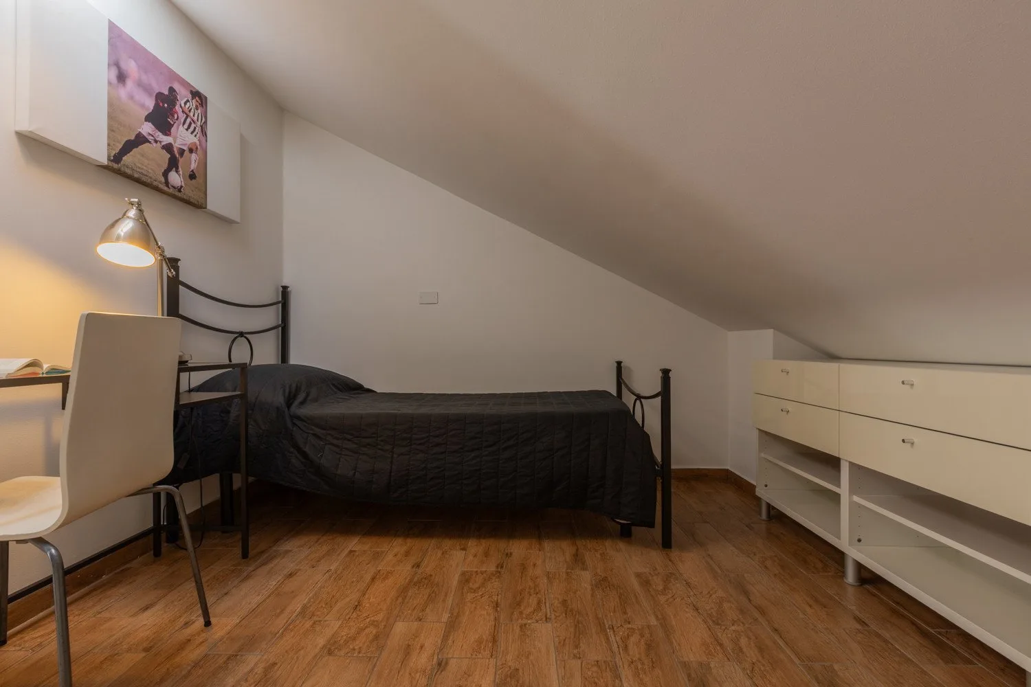 Un dormitorio pequeño con cama, escritorio y mueble bajo alargado bajo un techo inclinado.