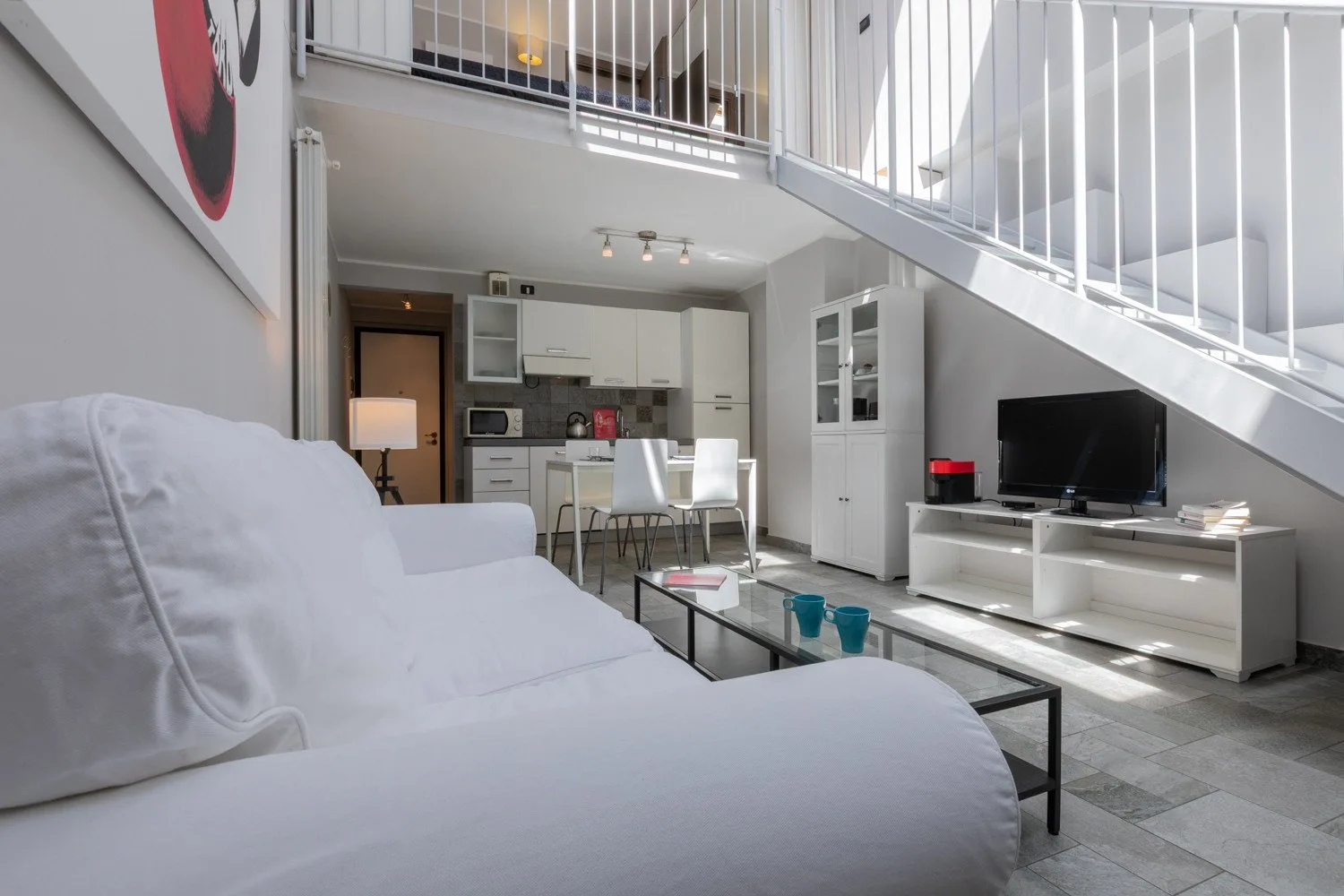 A bright loft living area with white seating, a glass coffee table, and a compact kitchen.