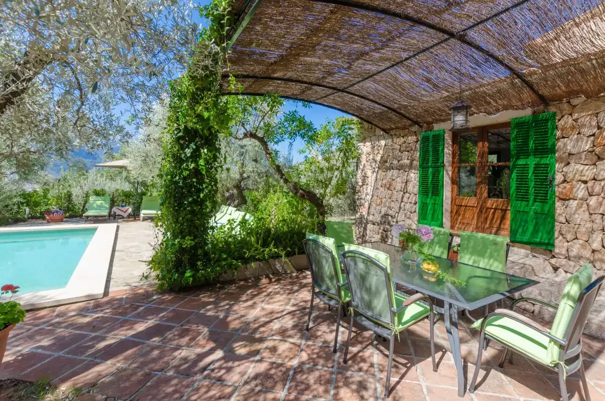 A shaded outdoor dining area beside a swimming pool and stone house.
