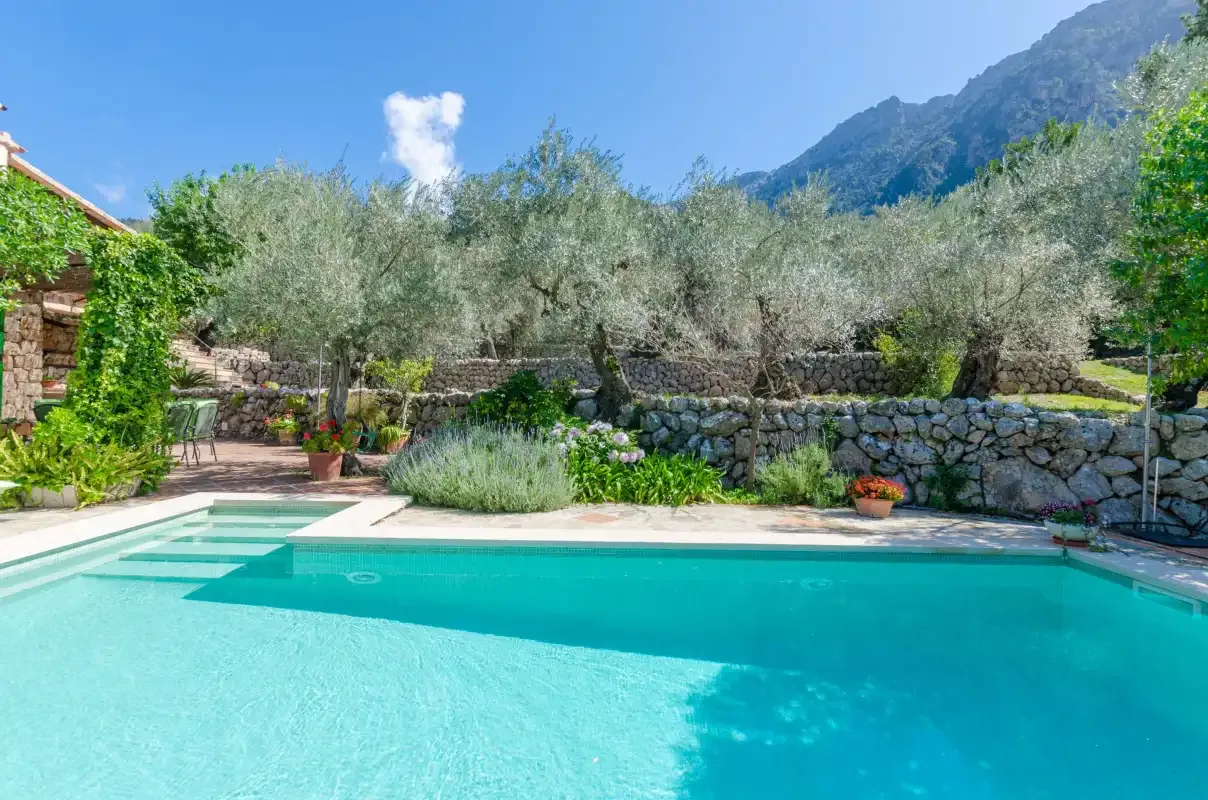 A swimming pool sits beside a stone garden wall, with trees and mountains beyond.