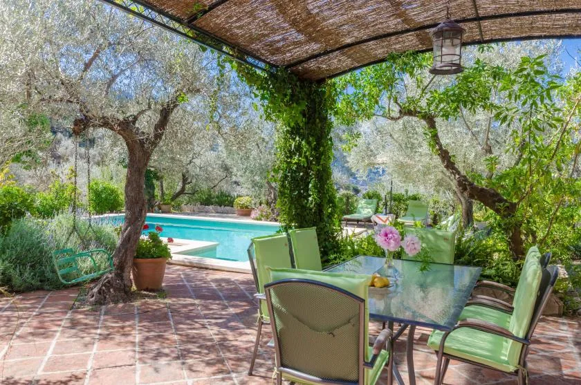 A shaded patio with outdoor seating overlooks a swimming pool and leafy garden.