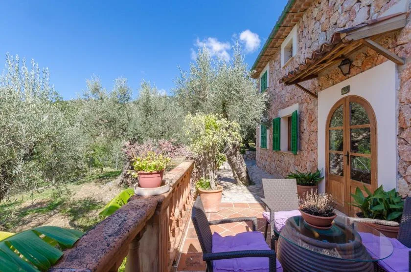 A stone house opens onto a sunny terrace with outdoor seating and potted plants.