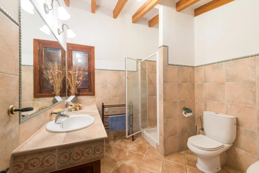A tiled bathroom with a sink, shower, and toilet beneath exposed wooden ceiling beams.