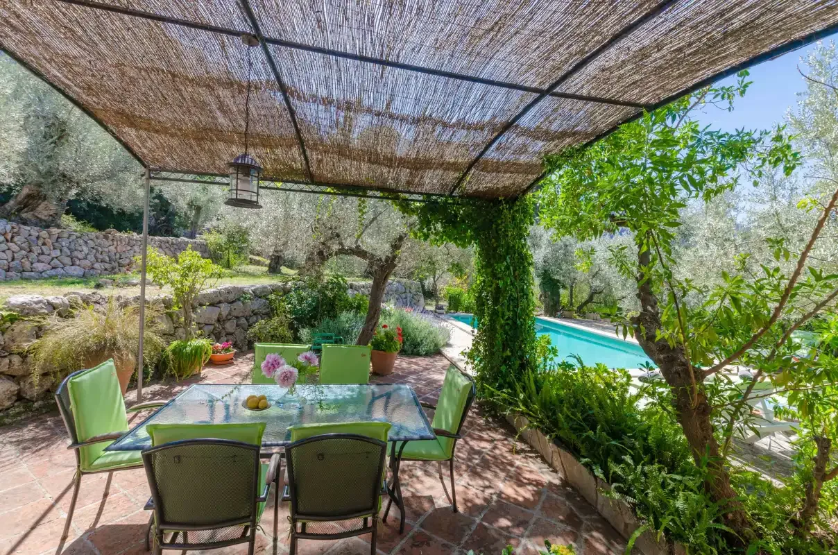 An outdoor dining area under a woven shade canopy overlooks a swimming pool.