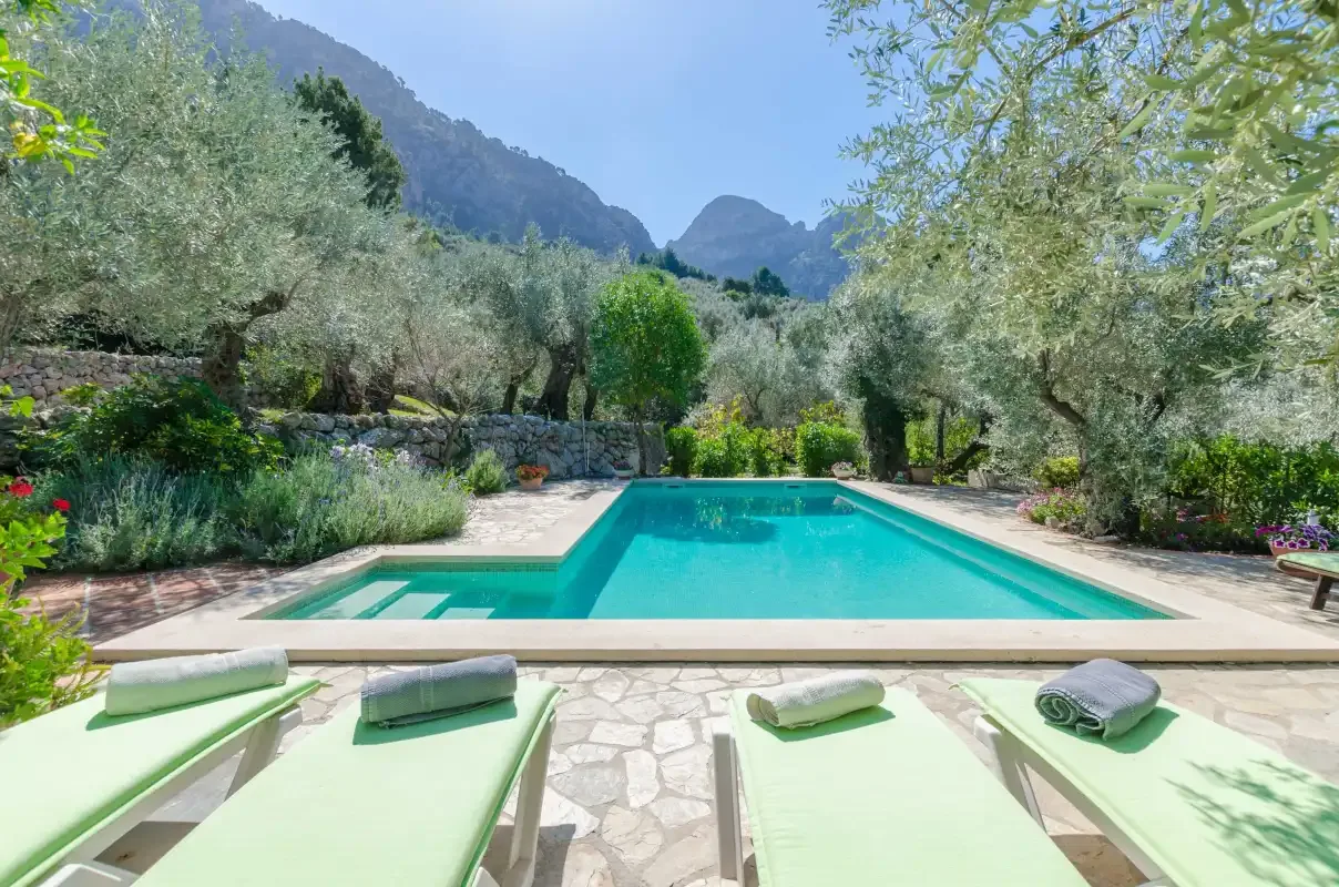 A swimming pool bordered by lounge chairs, greenery, and mountain views.