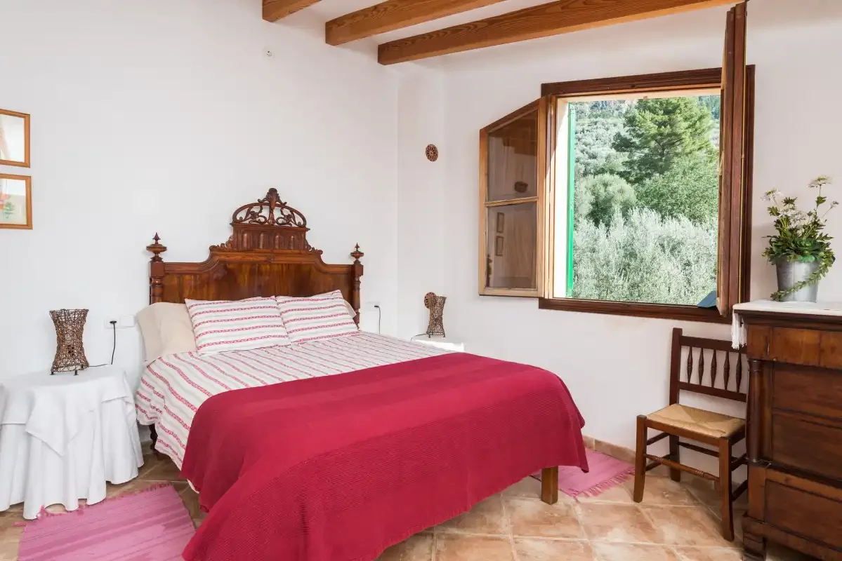 A bedroom with a bed covered by a red blanket beside an open window.
