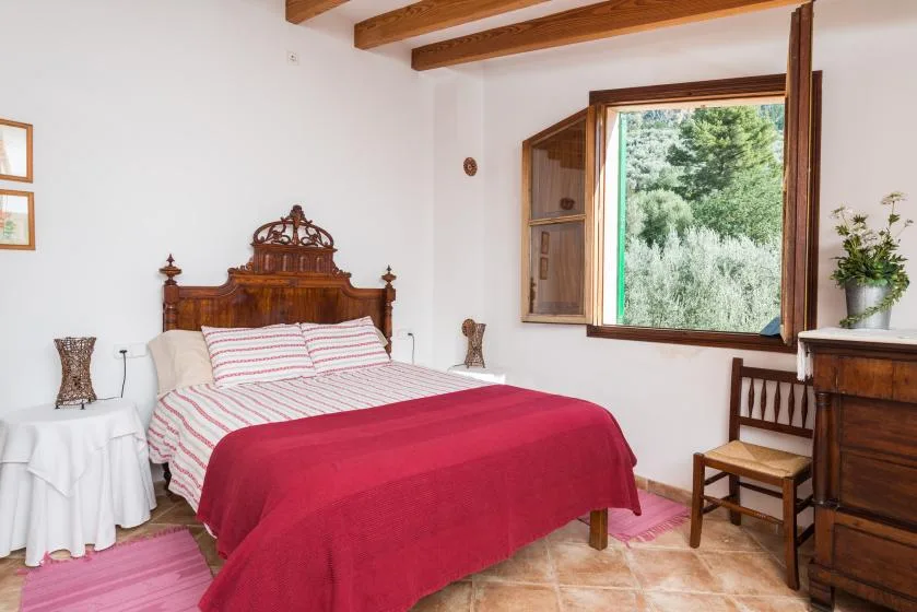 A bedroom with a bed covered by a red blanket beside an open window.