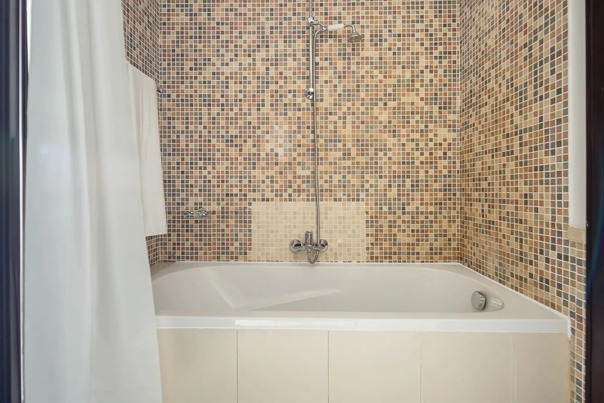 Una bañera con ducha está frente a una pared cubierta de pequeños mosaicos.