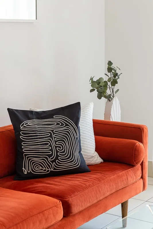 Un sofá naranja con cojines estampados está junto a un jarrón con vegetación.