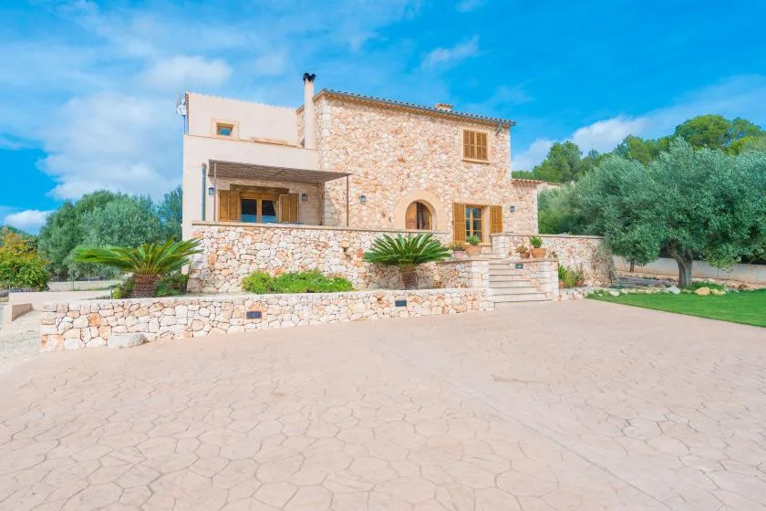 A stone house stands behind a paved courtyard with trees nearby.