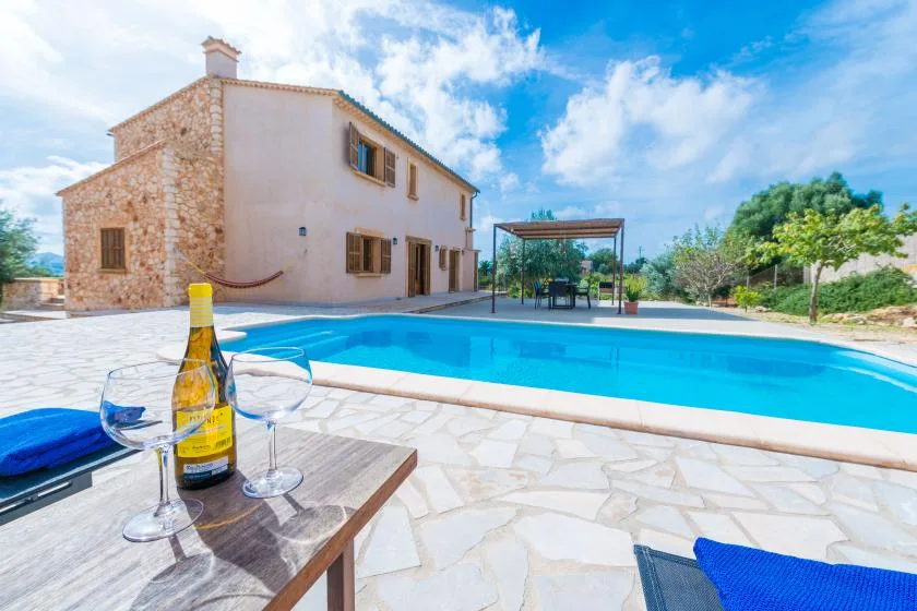 A house stands beside a swimming pool, with a table and glasses in the foreground.