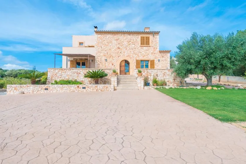 A stone house stands beyond a broad paved driveway, with trees and lawn nearby.