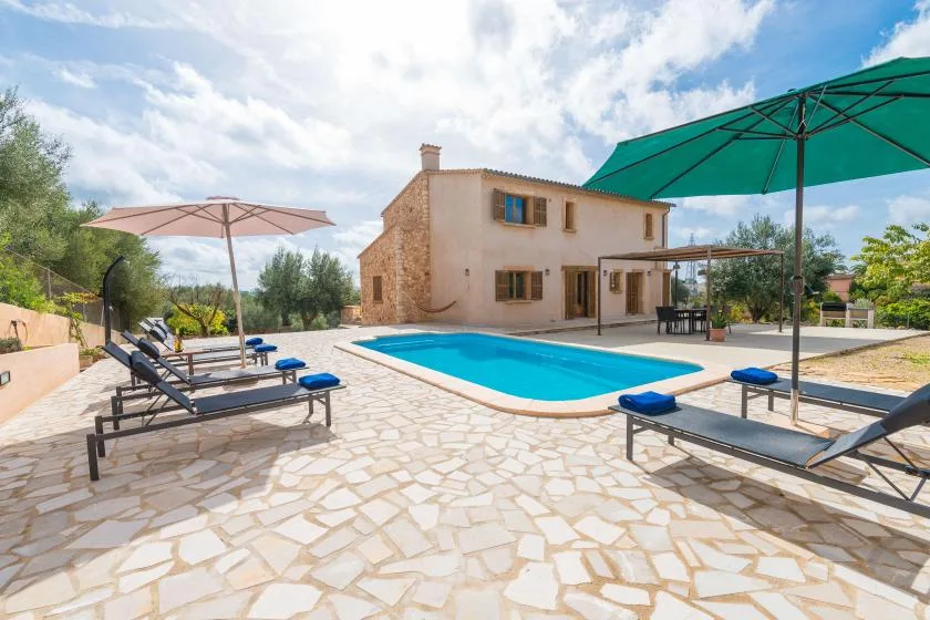 A swimming pool and sun loungers sit beside a two-story house on a paved patio.