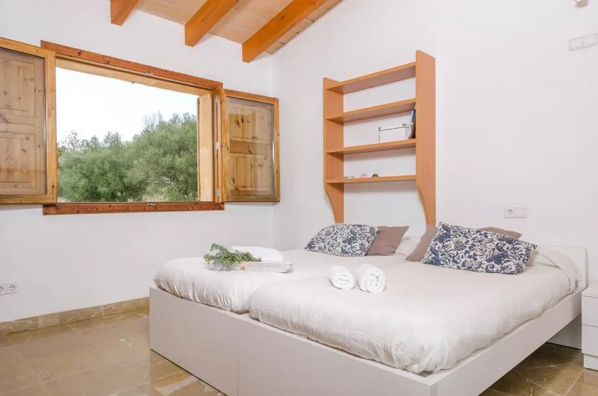 A bedroom with a bed beneath wooden ceiling beams and a window overlooking greenery.